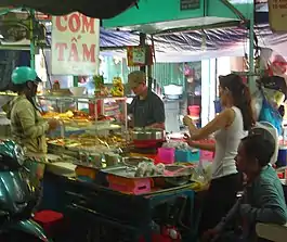 Com tam stall on the street. A board titled "Com Tam" is hung on top of the stall. On the stall, a cook is making com tam for his customers who are waiting around the stall for their to-go order. In front of him is a small, transparent glass cupboard where he keeps his ingredients ready to put in the dish. Behind the stall, it looks like a small building where this stall stores its stuff. Most of the time, in Vietnam, this "building" is the owner's house. This is how people do casual com tam business in Vietnam.