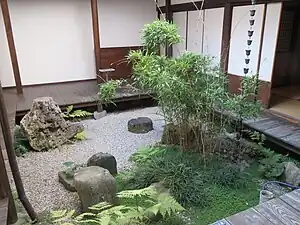A garden courtyard with an en about 30 centimetres (12 in) above the pale gravel, feature stones, a rain chain, and a planting of bamboo, ferns, grasses, and a creeping plant with small round leaves.