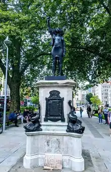 A black life-size statue of a black woman in contemporary clothes, standing with her right fist raised above her head. Statue is on a plinth with a placard placed at the base that reads "BLACK LIVES STILL MATTER". There is a background of an urban space and a tree.