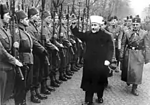 a man wearing Muslim mufti clothing holding his hand up in salute as he and a group of SS officers inspect a line of soldiers