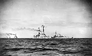 Several light gray warships are seen steaming in the distance; most are steaming generally toward the viewer, while the Königsberg-class cruiser is sailing in the opposite direction.