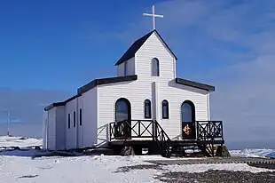 A white building with a black roof, a small porch, and a double height central tower topped with a white cross