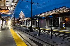 A low-angle shot of a side-platform two-track at-grade train station in the evening. In the background, an illuminated Italian Renaissance-style dome