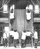 Students co-operate to scale a high wall. The activity involves lifting someone up, wall walking, and pulling someone up. c. 1900