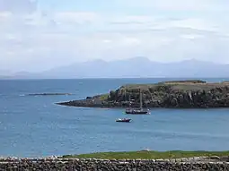 A two-masted yacht and a RIB lie at anchor on a sunny day off a grassy shore, with low cliffs beyond. A skerry lies further offshore to the left with high hills in the distance.