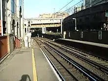A picture of the end of a side-platform light rail train station and the tracks leading away under an overpass. A third rail is visible in the foreground, ending at the end of the platform. An overhead catenary line is visible as well, extending as far as can be seen under the overpass.