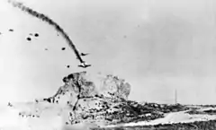 A blurry black and white photograph of two aircraft with numerous parachutes descending from them. One aircraft is trailing smoke and losing height