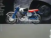 A pristine-looking 1960's-era single-cylinder motorcycle with a blue frame and bright red seat, and a Honda wing logo on the gas tank.