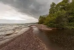 A stream of dark water flows from the right side of the images, away from the camera and some ways down a stony reddish beach, past a copse of green trees, before veering left across the beach and meeting a lake so wide that its far shores cannot be seen. The sky is cloudier on the right side, less so on left over the lake.