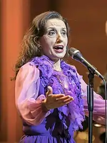 A woman with brown hair is wearing a purple blouse performing behind a microphone.