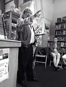 photo of a rotund bearded jolly man pointing to the audience with one hand while reading from the book of his poetry with his other.