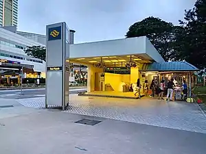 The rectangular-shaped station entrance with a connecting linkway (with a blue canopy) from the right