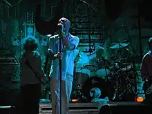 A blue-tinted photograph of musicians in front of an industrial background. From left to right: a long-haired male stands with his back to the camera playing bass guitar, a middle-aged Caucasian male sings into a microphone, a middle-aged Caucasian male plays behind a black-and-silver drum set on a riser, and a guitar player is mostly cropped from the extreme right of the photo.