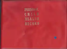 The front cover of a book, with a solid red background and the words 'PERSONAL CHILD HEALTH RECORD' in gold serif justified typeface on alternating lines. A small blue circular sticker is present in the bottom right.