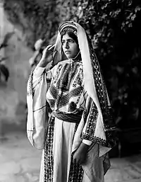 Image 5A young woman from Ramallah, c. 1898-1914. Until the 1940s, women of Palestine wore elaborate handcrafted garments. The creation and maintenance of these items played a significant role in their lives. A knowledgeable observer could determine a woman's village of origin and social status from her clothing. The circular band near this woman's forehead is a ring of coins made from a portion of her dowry money, and indicates that she is unmarried.'Ali Ra'if Efendi, (edited by Durova)