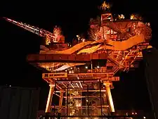 An offshore platform repurposed as an art installation, with walkways and foliage, illuminated by orange light at night.