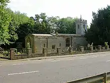 A stone church with a tower on the right. This has a battlemented parapet beyond which is a small pyramidal roof.