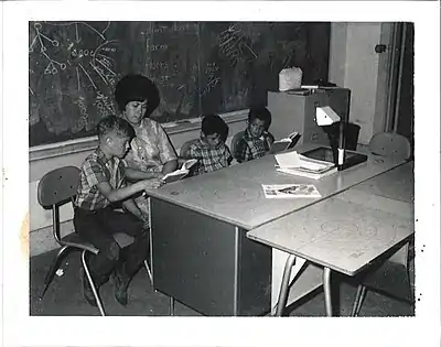 Three young boys sit at a table with their teacher.  The teacher points to the words that one boy is reading in his textbook.