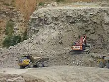 A photograph showing quarrying operations with a mechanical digger in the centre extracting rock from a cliff face. To the left, a lorry is just leaving with a full load of rock, and to the right, another lorry is waiting to collect its load from the digger.