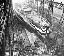 A large warship, still missing most of its superstructure, sits in a dry dock, awaiting its launch. The ship is draped in a large banner and surrounded by crowds of spectators; a huge gantry towers over the ship.