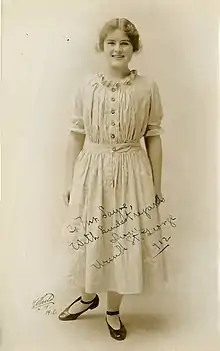 A young white woman standing, smiling, wearing a light-colored dress with buttons down the bodice front and a ruffled neckline.